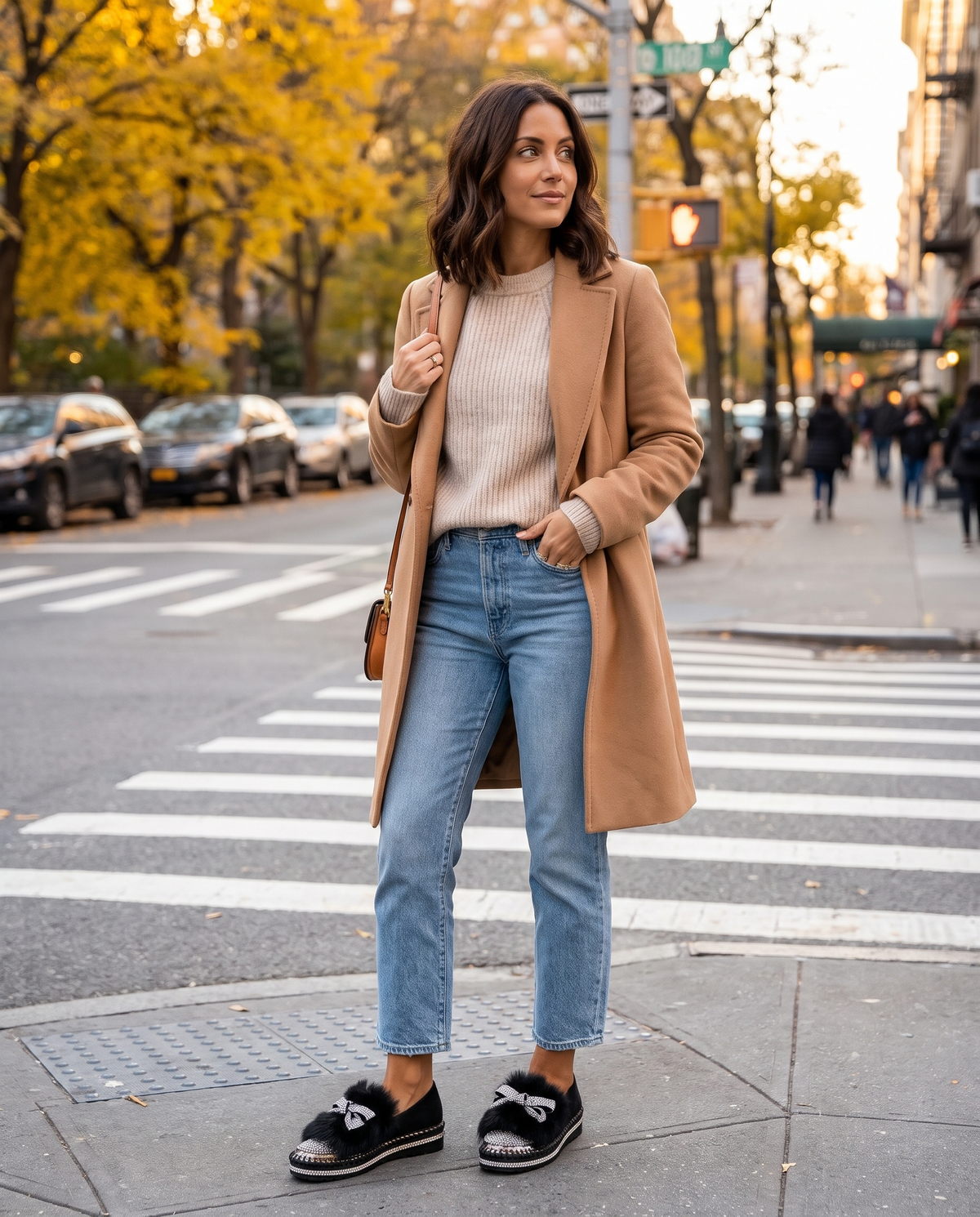Woman in a camel coat and cropped jeans wearing black crystal bow loafers at a fall city crosswalk